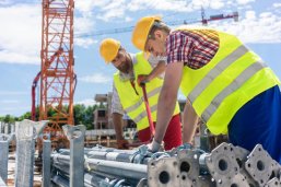 Zwei Bauarbeiter in Warnwesten und Schutzhelmen, die auf einer Baustelle Metallteile vorbereiten.