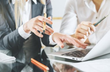 Zu sehen sind die Hände und Arme von zwei Personen in Business-Kleidung, während sie an einem Tisch sitzen. Eine Person deutet auf einen geöffneten Laptop, während beide Personen Kugelschreiber in den Händen halten.