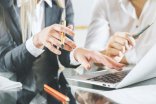 Zu sehen sind die Hände und Arme von zwei Personen in Business-Kleidung, während sie an einem Tisch sitzen. Eine Person deutet auf einen geöffneten Laptop, während beide Personen Kugelschreiber in den Händen halten.