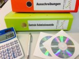 Ein Ordner mit der Beschriftung "Ausschreibungen", ein Ordner mit der Beschriftung "Zentrale Submissionsstelle", ein Taschenrechner, ein Kugelschreiber und zwei CDs liegen auf einem Tisch.