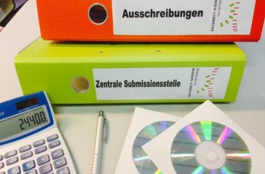 Ein Ordner mit der Beschriftung "Ausschreibungen", ein Ordner mit der Beschriftung "Zentrale Submissionsstelle", ein Taschenrechner, ein Kugelschreiber und zwei CDs liegen auf einem Tisch.