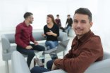 Junger Mann schaut in die Kamera im Hintergrund ein Team bei der Arbeit am Tisch