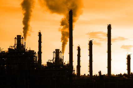 Heller Hintergrund, im Vordergrund diverse schwarze  Industrieschornsteine und Industrieanlagen, aus denen Dampf aufsteigt.