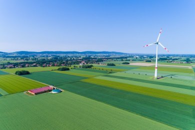 Windkraftanlage Landwirtschaftliche Felder, inmitten der sich ein Betriebsgelände befindet und ein großes Windrad auf den Feldern steht.