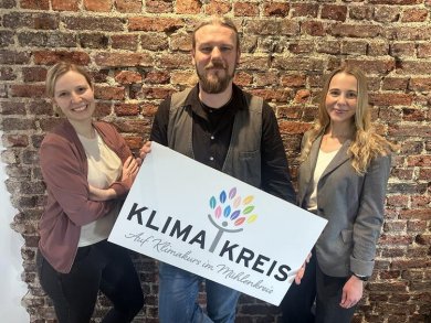 Klimakreis Team Gruppenfoto von dem Klimaschutz- und Klimaanpassungsteam im Kreis Minden-Lübbecke. Zwei Frauen und ein Mann; halten gemeinsam ein Schild mit dem Logo des Klimakreises vor sich.