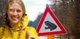 Junge blonde Frau im Fokus des Bildes , die eine gelbe Jacke trägt und den hochgestreckten Daumen in die Kamera richtet. Im Hintergrund befindet sich das Verkehrszeichen zur Warnung vor Amphibienwanderung, auf dem ein Frosch abgebildet ist