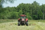 Roter fahrender Traktor auf einer grünen Wiese, der mit einem Heuwender das Gras bearbeitet