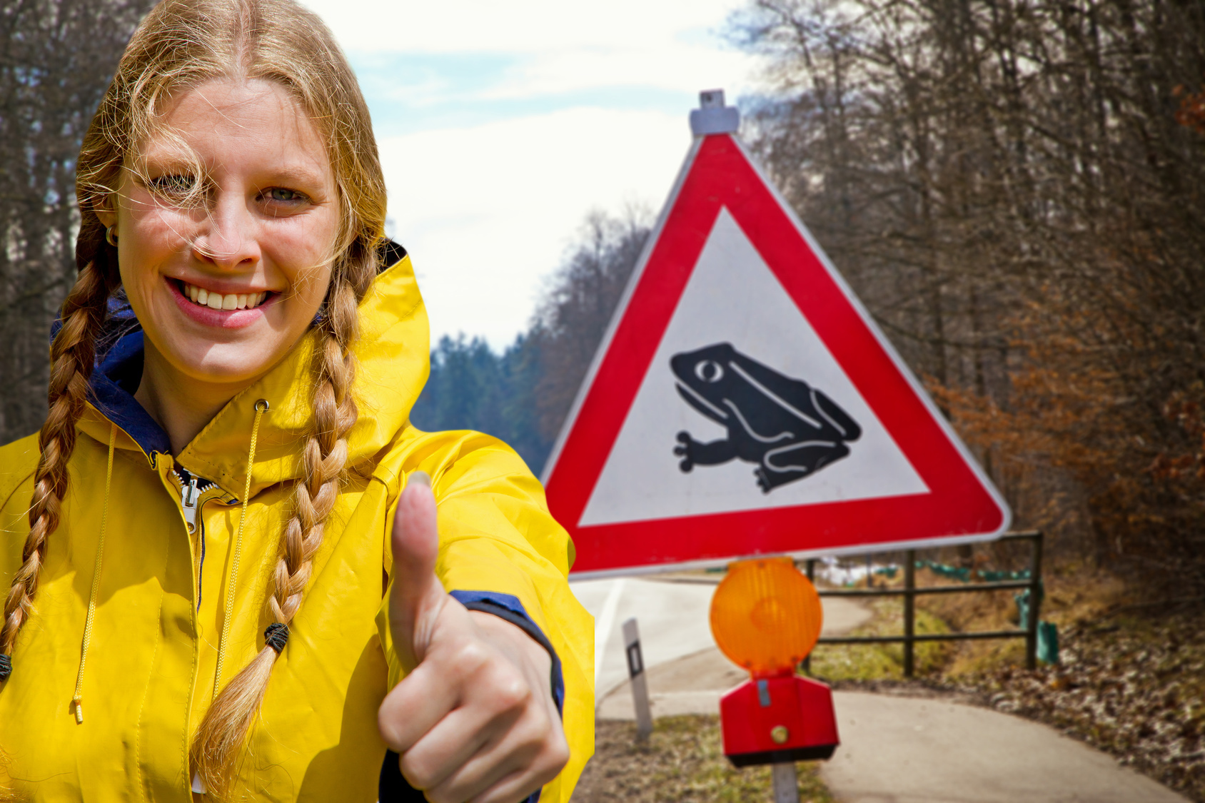 Junge blonde Frau im Fokus des Bildes , die eine gelbe Jacke trägt und den hochgestreckten Daumen in die Kamera richtet. Im Hintergrund befindet sich das Verkehrszeichen zur Warnung vor Amphibienwanderung, auf dem ein Frosch abgebildet ist